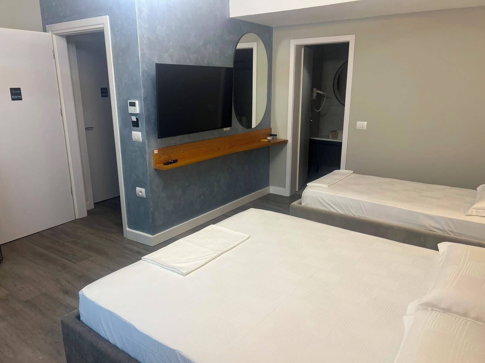 Habitación de hotel con dos camas, un televisor montado en la pared, un espejo y un estante de madera.