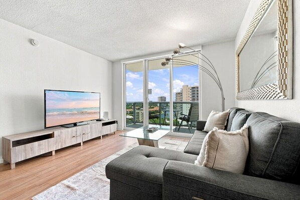 A modern living room with a flat-screen TV, a grey sofa, and a glass coffee table. - Spacious & Modern apt for a family beach vacation (Hollywood)