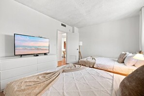 Ein Schlafzimmer mit einem Fernseher auf einer weißen Kommode, einem Bett mit einer beige farbenen Decke und einer Nachttischlampe.