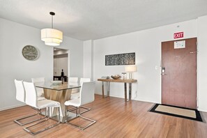 Ein modernes Interieur mit einem Esstisch aus Glas, weißen Stühlen, Holzboden, einer Pendelleuchte, einer Wanduhr, einer Dekoration und einer Ausgangstür.