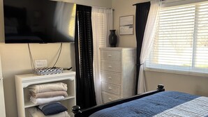 Ein Schlafzimmer mit einem Bett, einem an der Wand montierten Fernseher, einem Schminktisch und einem Fenster mit Vorhängen.