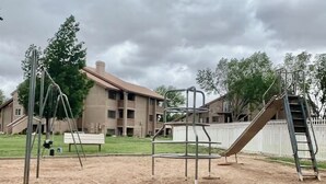 Ein Spielplatz mit Schaukeln und Rutsche, umgeben von einer Rasenfläche und einem Gebäude im Hintergrund.