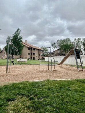 A playground with swings and a slide, surrounded by a grassy area and a building in the background. - Sunny Getaway at Sports Village (St. George)