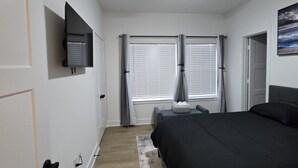 Un dormitorio con una cama, ventilador de techo, televisor montado en la pared y una ventana con persianas.