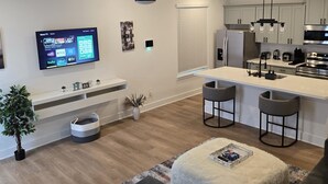 Una sala de estar moderna con un sofá gris, un televisor de pantalla plana montado en la pared, un ventilador de techo y una zona de cocina con refrigerador, microondas y taburetes de bar.