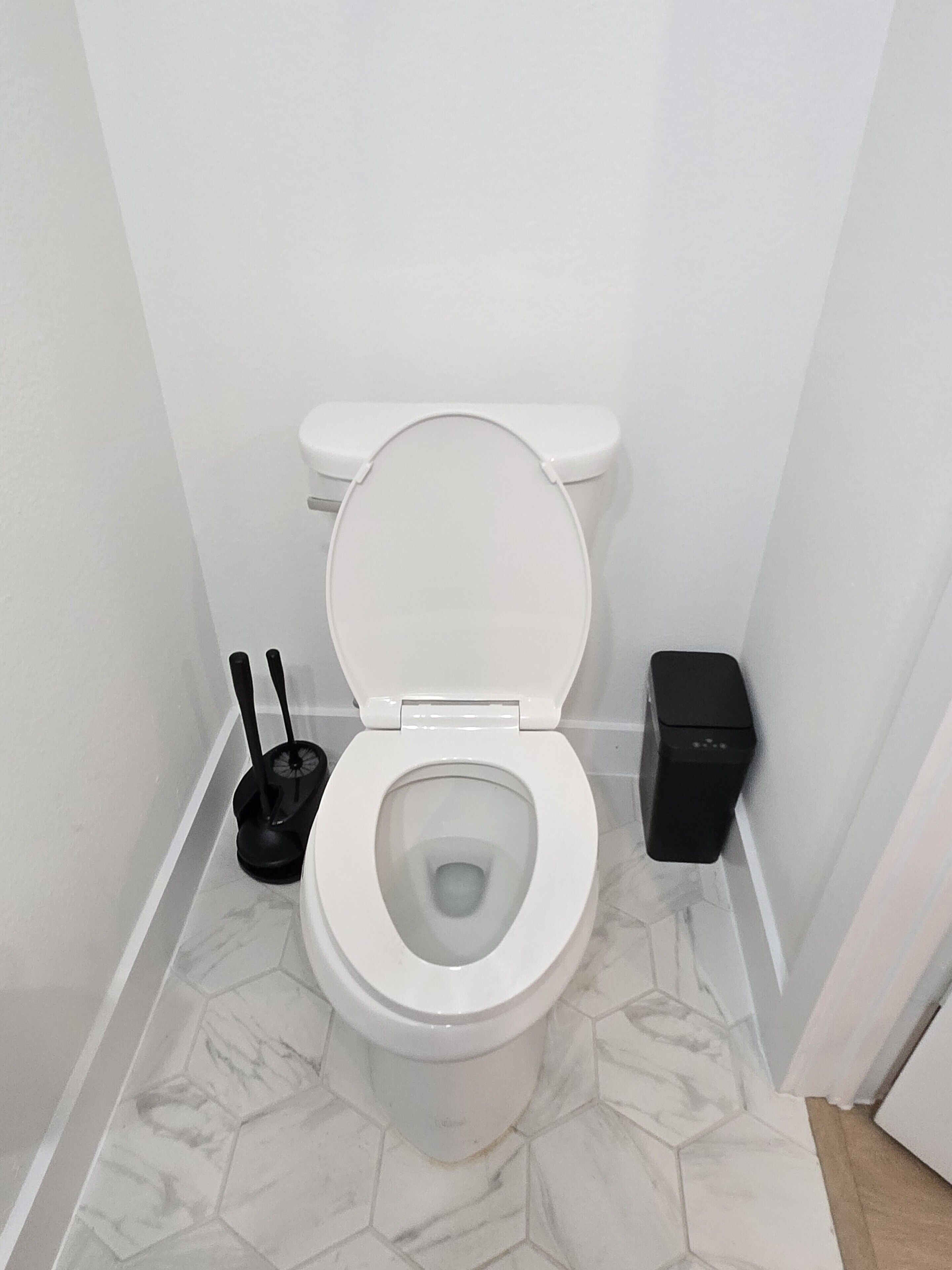 Un inodoro blanco con la tapa baja, un bidé negro y un cubo de basura en un baño con suelo de mármol hexagonal.