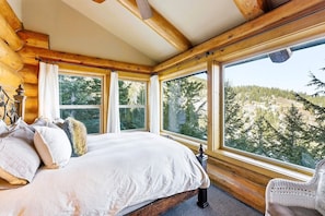 Ein Schlafzimmer einer Holzhütte mit einem Bett, einem Sessel und großen Fenstern mit Blick auf Bäume und Hügel.