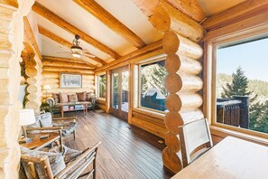 Ein Interieur einer Blockhütte mit Holzbalkendecke, Deckenventilator, einer Couch, einem Couchtisch und einem großen Fenster mit Blick ins Freie.