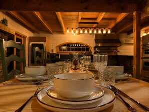 A dining area with a wooden table set for two, a large bowl in the center, and a kitchen in the background.