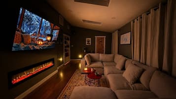 A cozy living room with a large flat-screen TV, a fireplace, a sectional sofa, and a red coffee table.