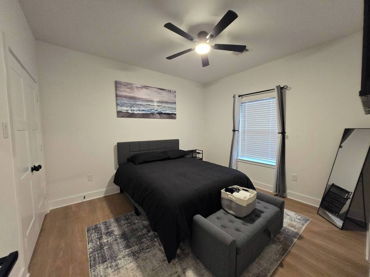 A bedroom with a bed, a ceiling fan, a window with blinds, a bedside table with a lamp, and a wall-mounted mirror.