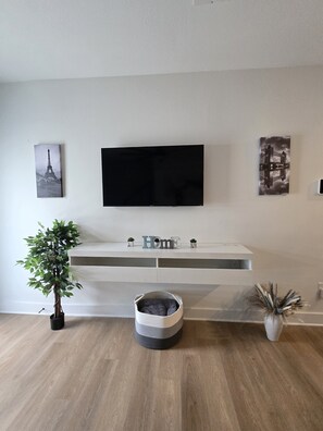 A modern living room with a flat-screen TV mounted on the wall, a potted plant, a decorative basket, and a shelf with small plants and the word 'HOME' on it. - NRG Stadium Stay | Texans Football | Suite (Houston)