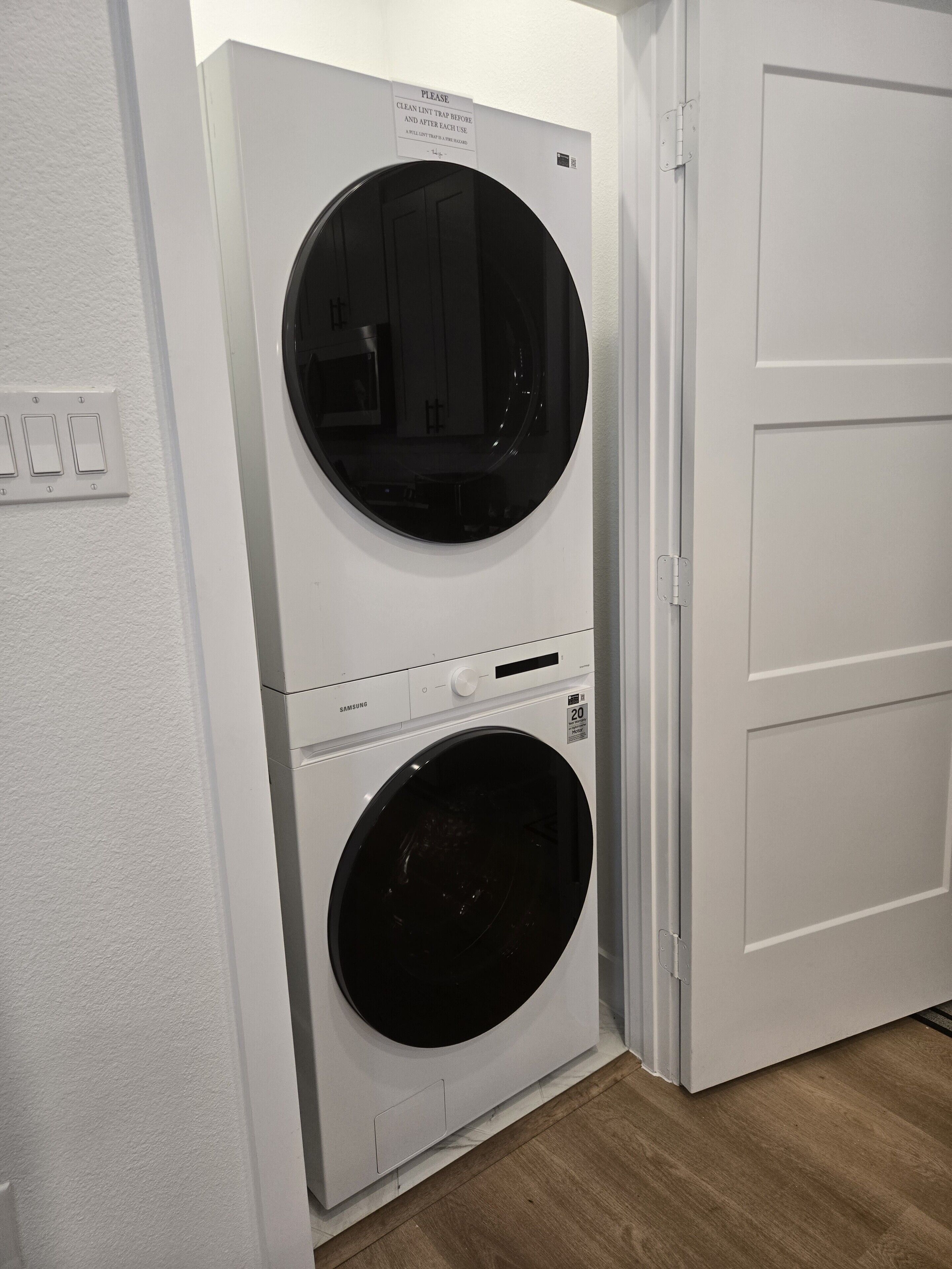A front-loading Samsung washer and dryer unit installed in a corner of a room with white walls and a wooden floor.