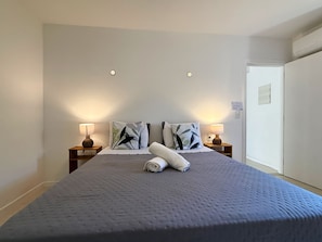 Una cama bien hecha con colcha gris, dos lámparas de noche y dos almohadas con motivos de hojas.