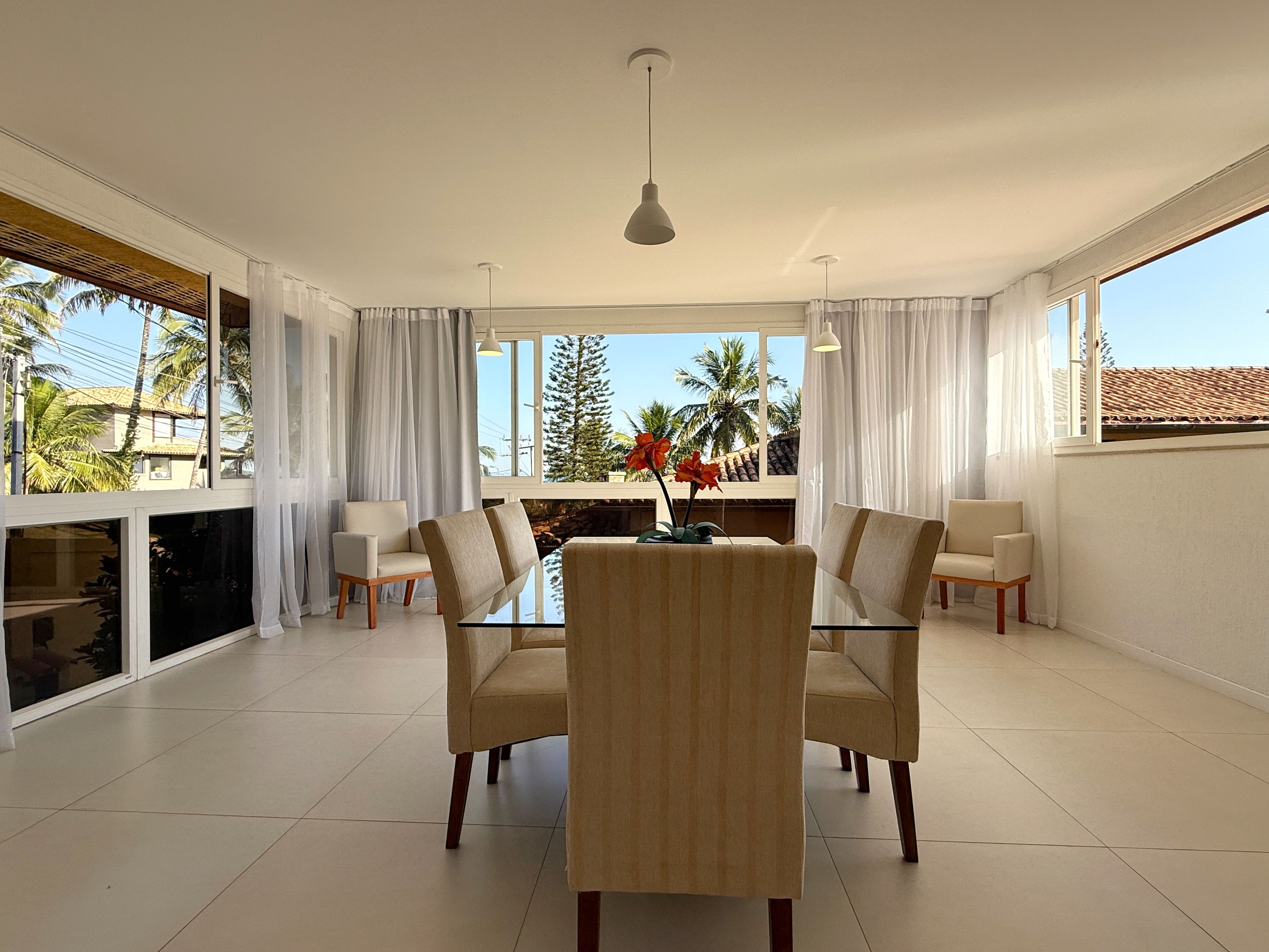 A modern dining room with a glass table and chairs, large windows with curtains, and a view of palm trees.