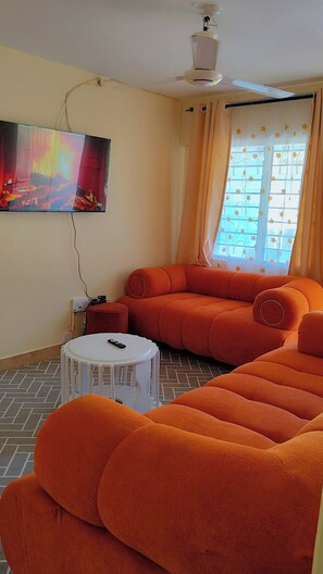 A living room with a sofa, a television mounted on the wall, a ceiling fan, and a window with curtains. - Comfortable and safe place not to far from the beach and it is family friendly. (Mombasa)