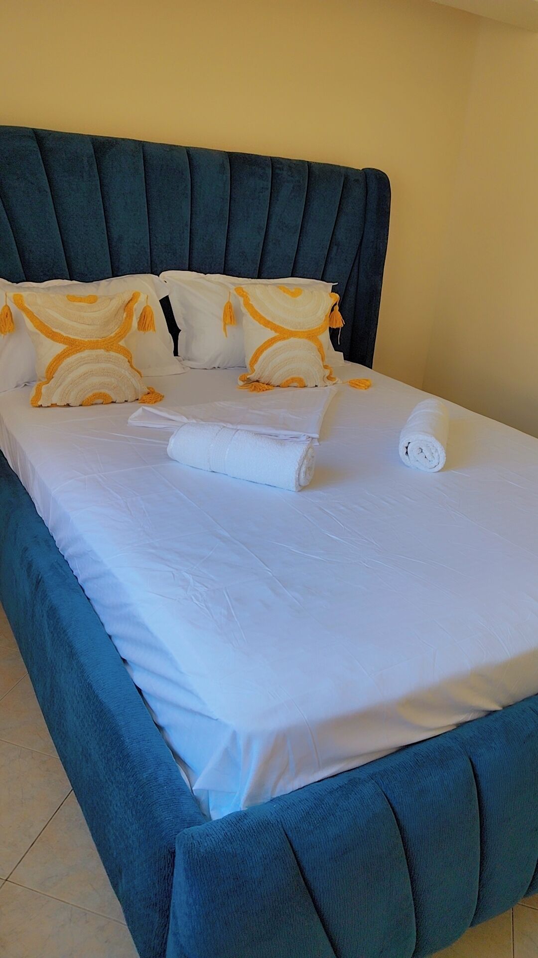 Una cama con cabecera tapizada en azul, ropa de cama blanca y dos almohadas decorativas con flecos.