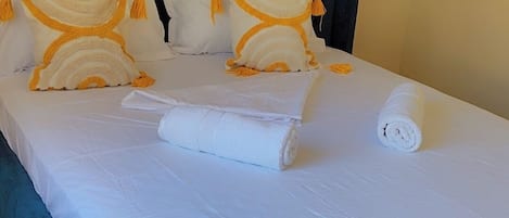 Una cama con cabecera tapizada en azul, ropa de cama blanca y dos almohadas decorativas con flecos.