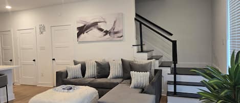 A modern living room with a gray sectional sofa, a white ottoman, a ceiling fan, and a staircase with black railings.