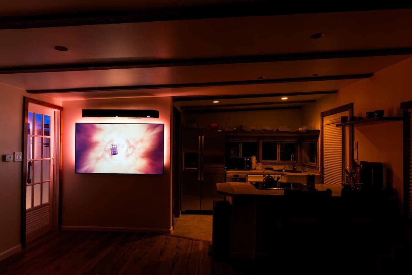A dimly lit room with a flat-screen TV mounted on the wall, a wooden floor, and a kitchen area in the background.
