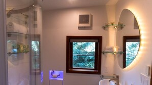 A bathroom with a glass shower enclosure, a white sink, a mirror with a curved frame, and a window with a view of trees. - Blue River Studio Hideaway (Breckenridge)
