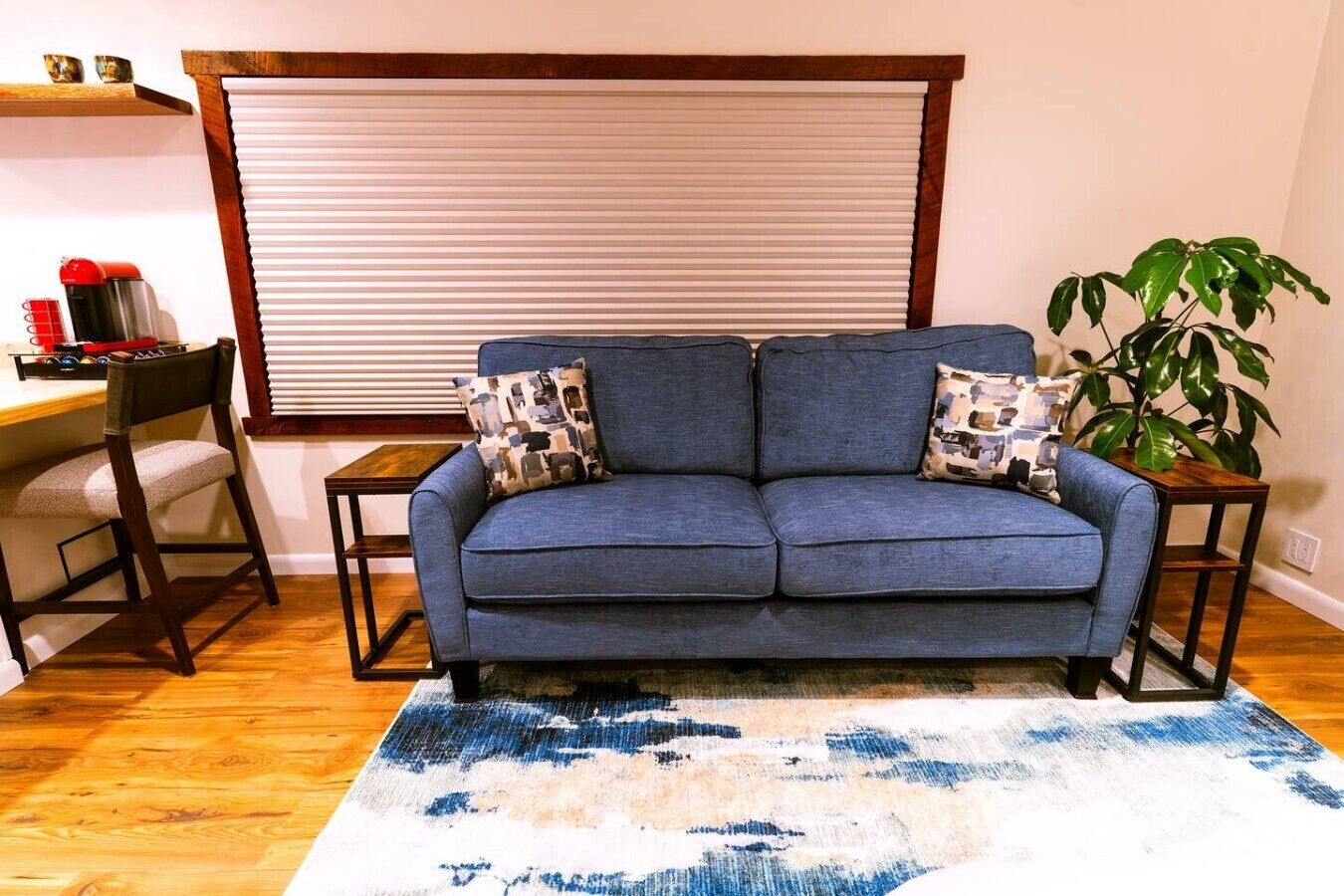 A living room with a sofa, two side tables, a coffee machine, a chair, a plant, and a window with blinds.