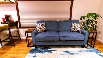 A living room with a sofa, two side tables, a coffee machine, a chair, a plant, and a window with blinds.