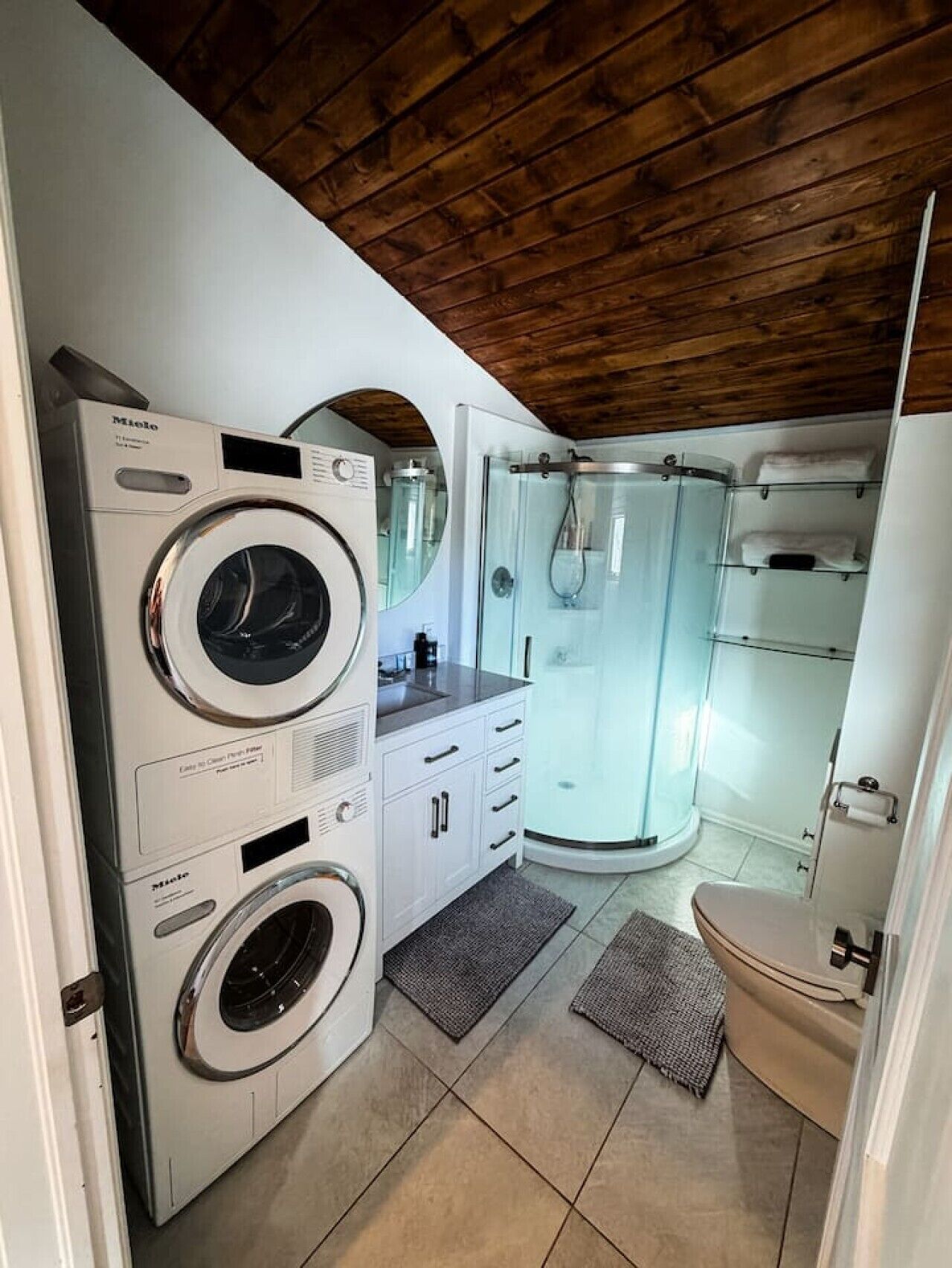A compact laundry room with a front-loading washing machine, a dryer, and a glass-enclosed shower.