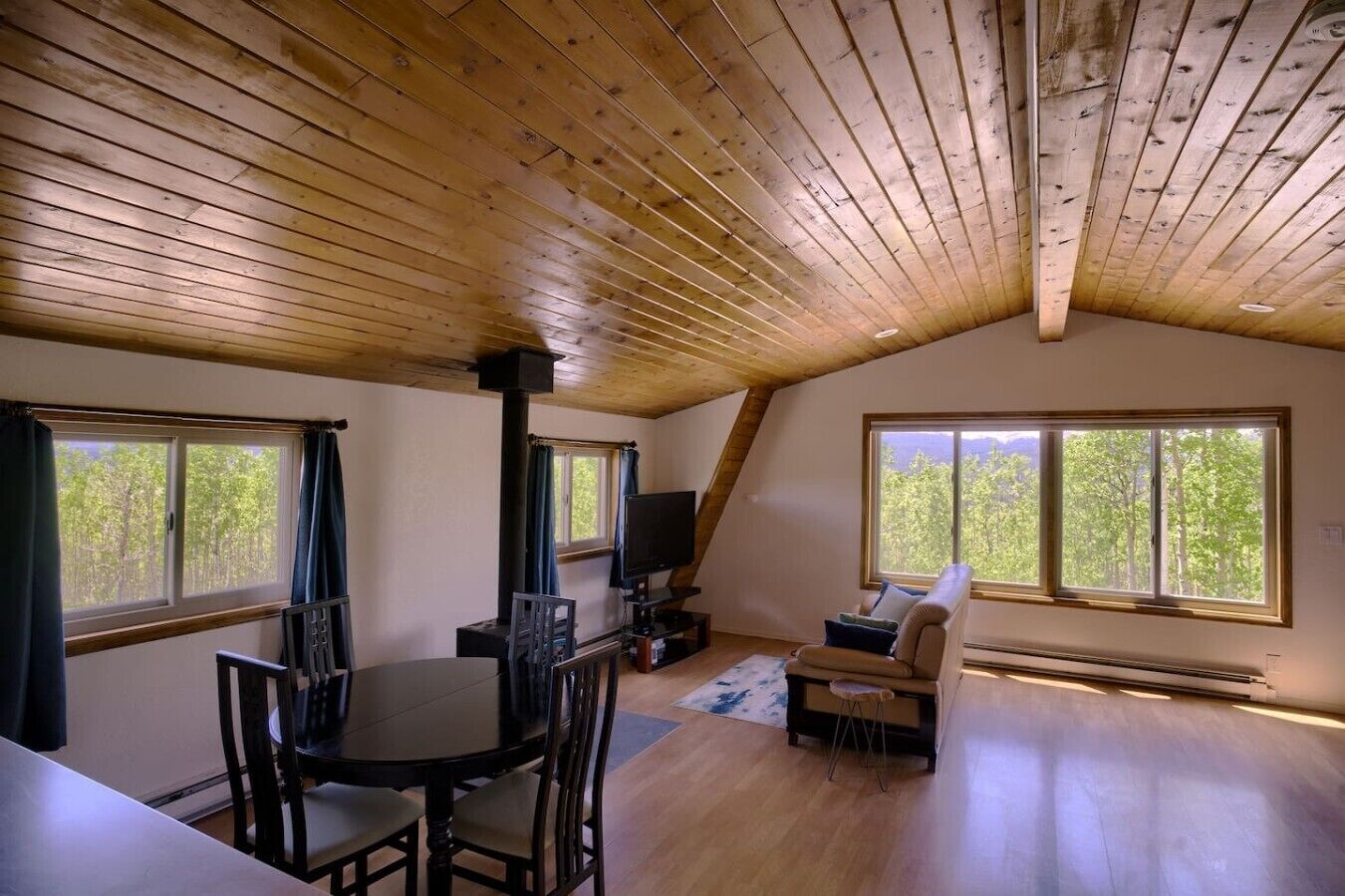 A cozy room with a dining table, chairs, a sofa, and a TV. There are large windows providing natural light and a view of trees.