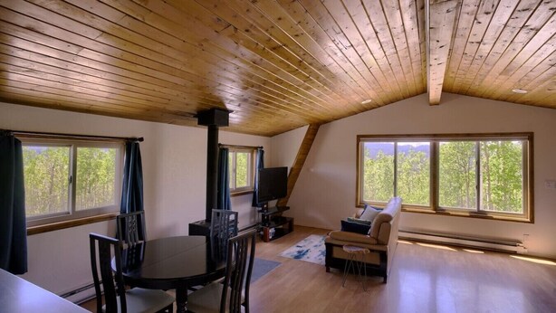 A cozy room with a dining table, chairs, a sofa, and a TV. There are large windows providing natural light and a view of trees.