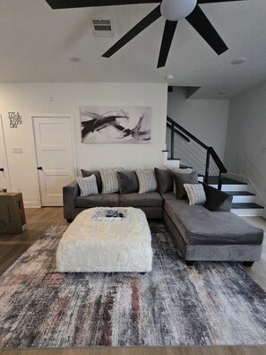 A modern living room with a sectional sofa, a white fluffy ottoman, a large abstract painting, and a staircase. - Texans Season | NRG Stadium | Houston 2BR (Houston)