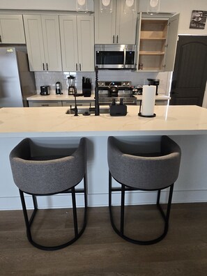 A modern kitchen with white cabinets, a central island with bar stools, and a coffee machine. - Texans Season | NRG Stadium | Houston 2BR (Houston)