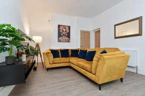 Ein Wohnzimmer mit einem senffarbenen Sofa, einer Stehlampe, einem Topfpflanze und einem gerahmten Bild an der Wand.