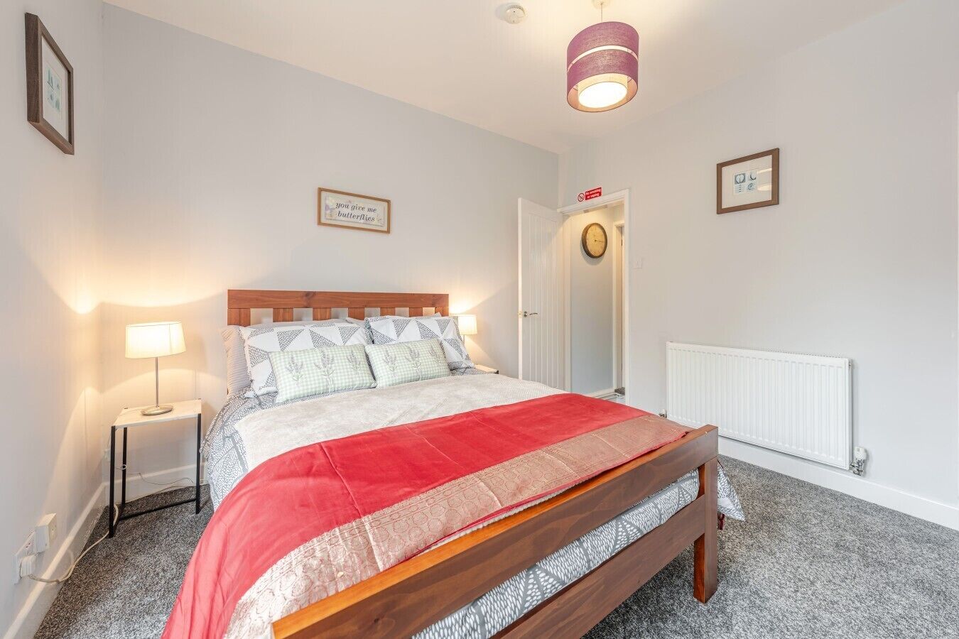 Een slaapkamer met een houten bed, een rood-wit dekbedovertrek, een nachtkastje met een lamp en een witte radiator.