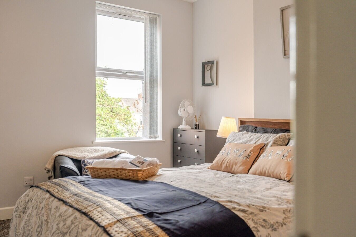 A bedroom with a large window, a bedside table with a lamp, and a bed with patterned bedding.