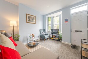 Ein Wohnzimmer mit Sofa, Sessel, kleinem Tisch und Fernseher. Eine Tür mit einem Schild darauf und ein Fenster mit Vorhängen.