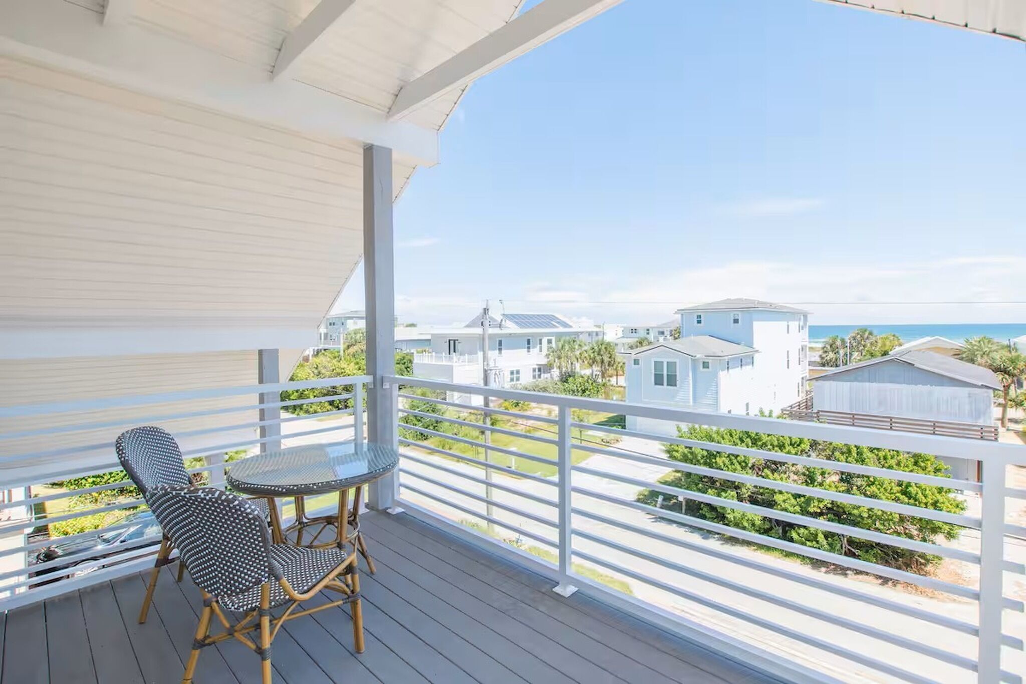 Un balcon abrité, doté d’une table et de chaises, qui offre une vue sur un quartier résidentiel avec ses maisons et une plage.