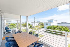 Une terrasse extérieure couverte, avec une table en bois et des chaises bleues, donnant sur un quartier résidentiel avec des maisons et de la verdure.