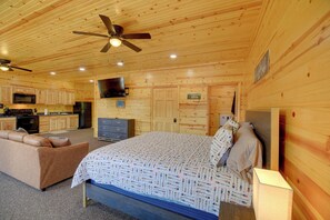 Une pièce aménagée dans le style d’un chalet en rondins, avec un lit, un canapé, une télévision, un coin cuisine et un ventilateur de plafond.
