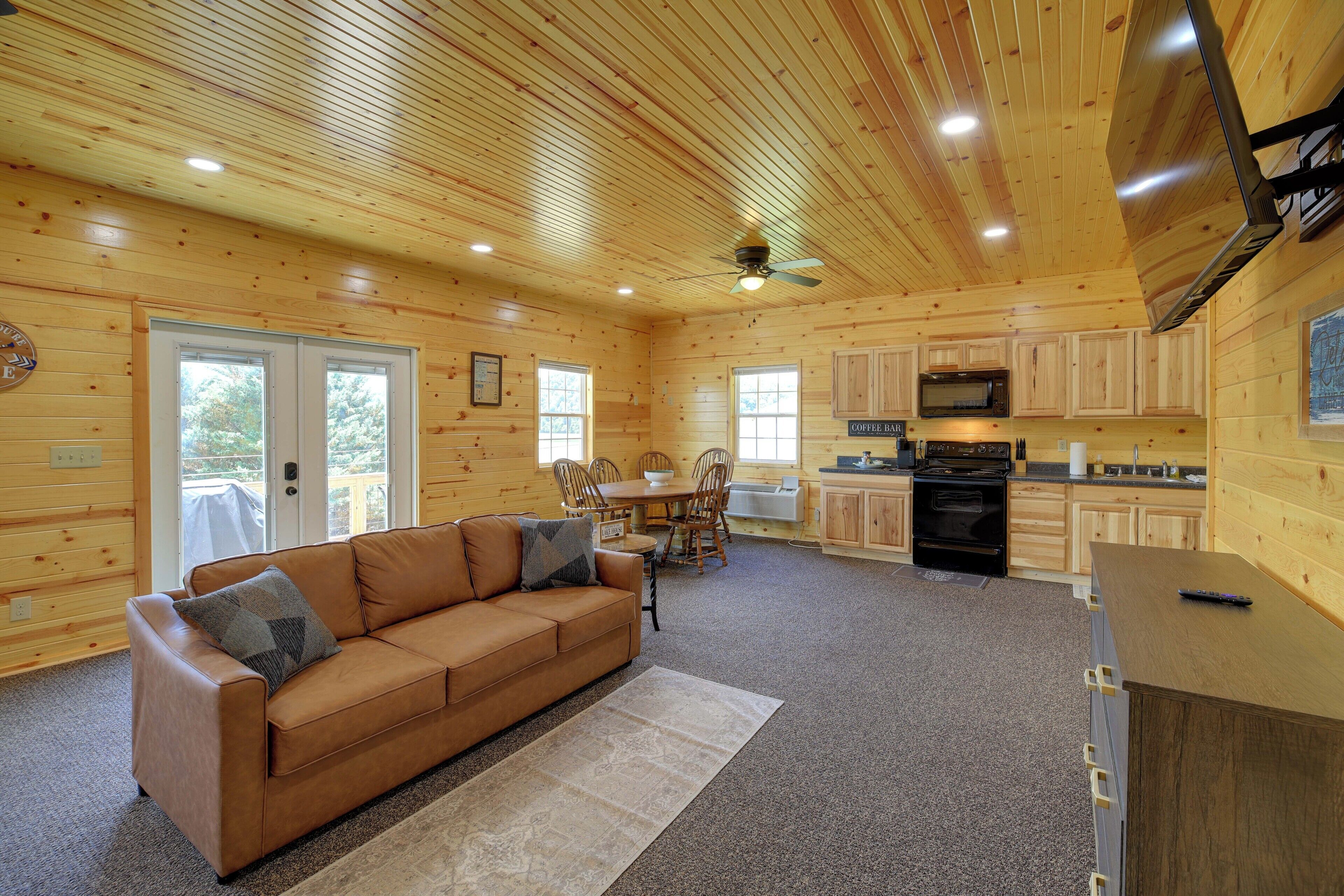 A cabin interior with a wooden interior, a sofa, a dining table, chairs, a kitchen area, and a ceiling fan.