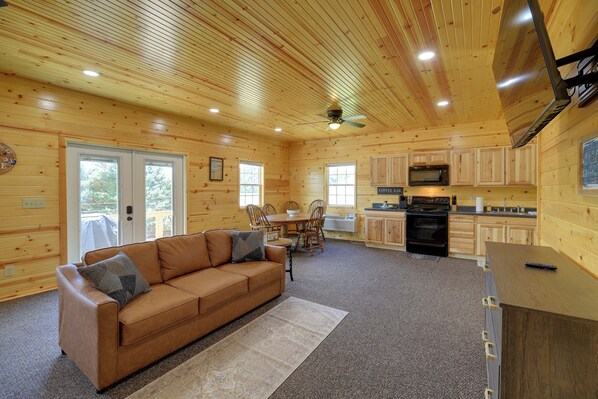 A cabin interior with a wooden interior, a sofa, a dining table, chairs, a kitchen area, and a ceiling fan.