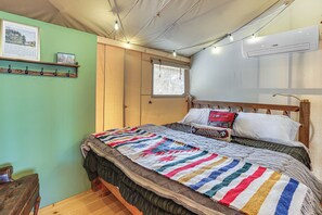 A bedroom with a bed, a wooden headboard, a window with curtains, a wall-mounted air conditioner, and a shelf with a framed picture.