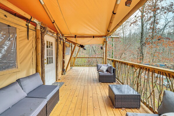 A covered wooden deck with wicker furniture, a view of trees, and a wooden railing.