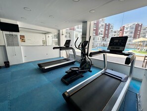 A gym with treadmills, an elliptical, and a rowing machine, with a view of a swimming pool and buildings outside. - Modern Pool-View Studio with Gym, Pool Pub & Restaurant in Aqua Palms Resort (Hurghada)