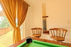 Ein Billardtisch mit Spielstöcken, zwei Holzstühle und ein Fenster mit Vorhängen.