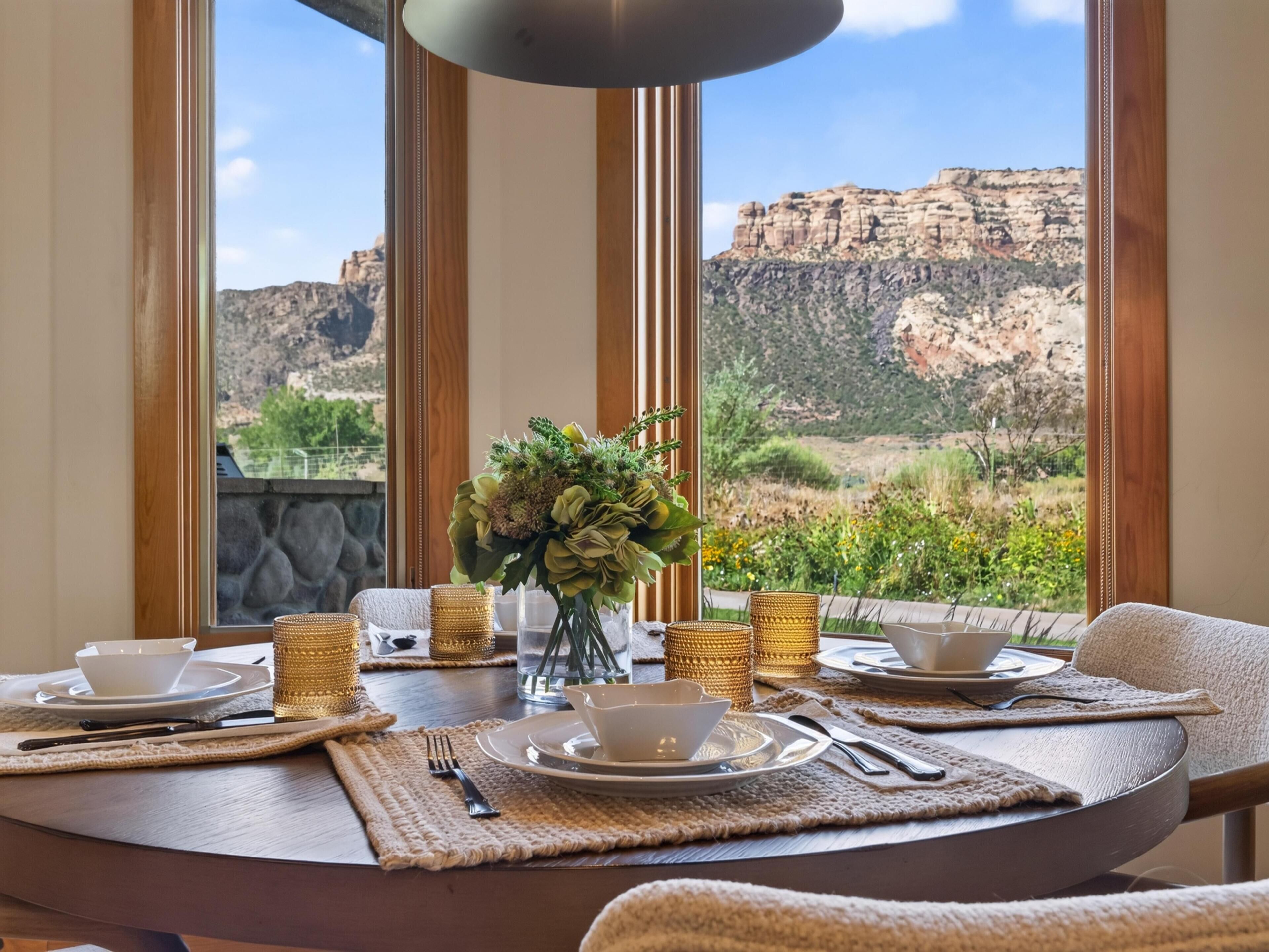 A dining table set with plates, cups, and cutlery, with a view of a mountainous landscape through large windows.