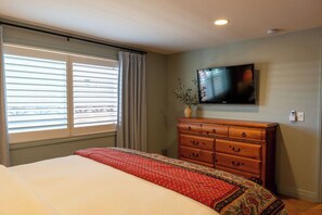A bedroom with a bed, a dresser, a television mounted on the wall, and a window with blinds.