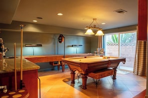 A pool table in a room with a clock on the wall, a bar area, and a window with curtains.
