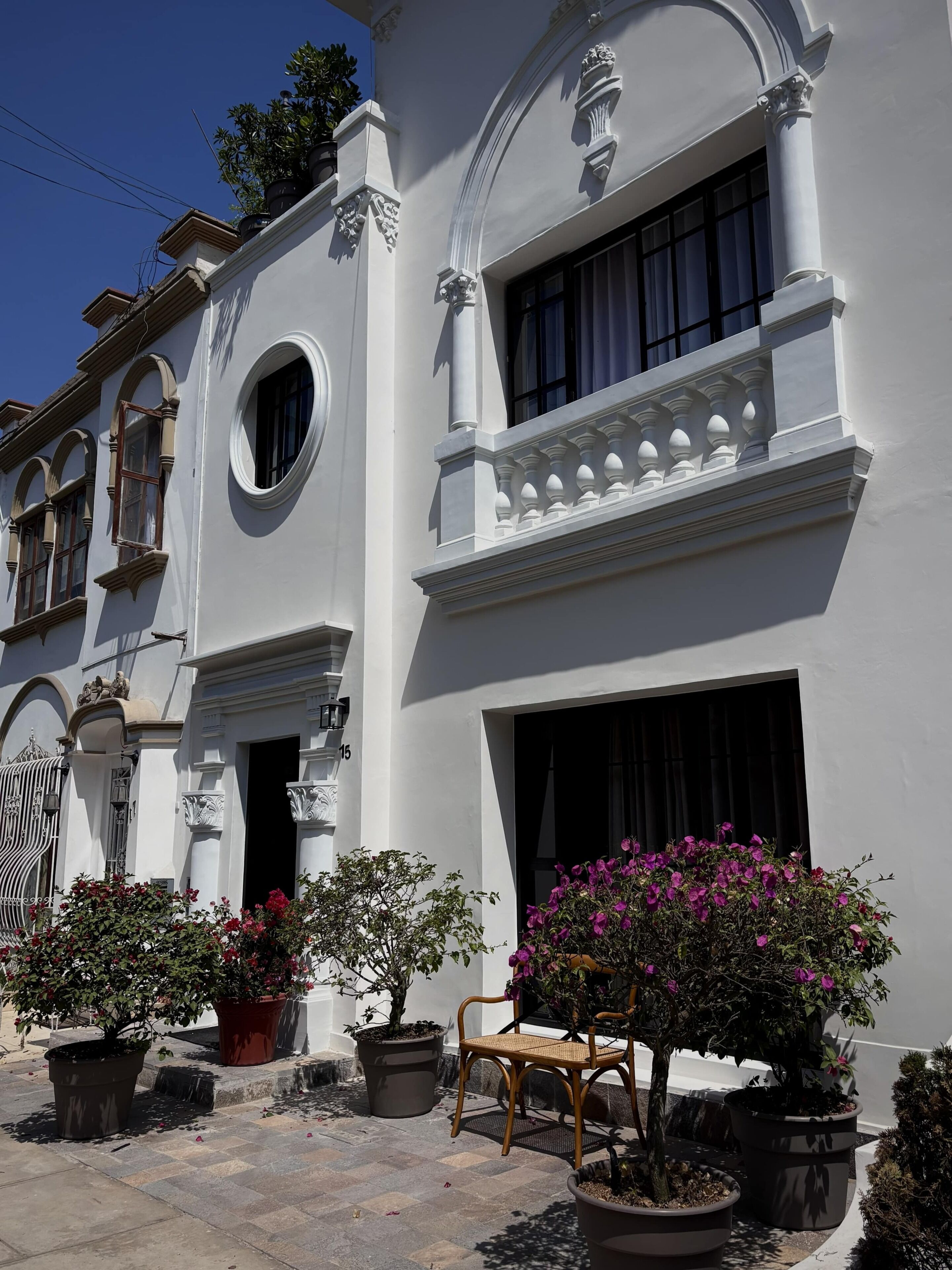 A white building with a balcony, potted plants, and a wooden chair.
