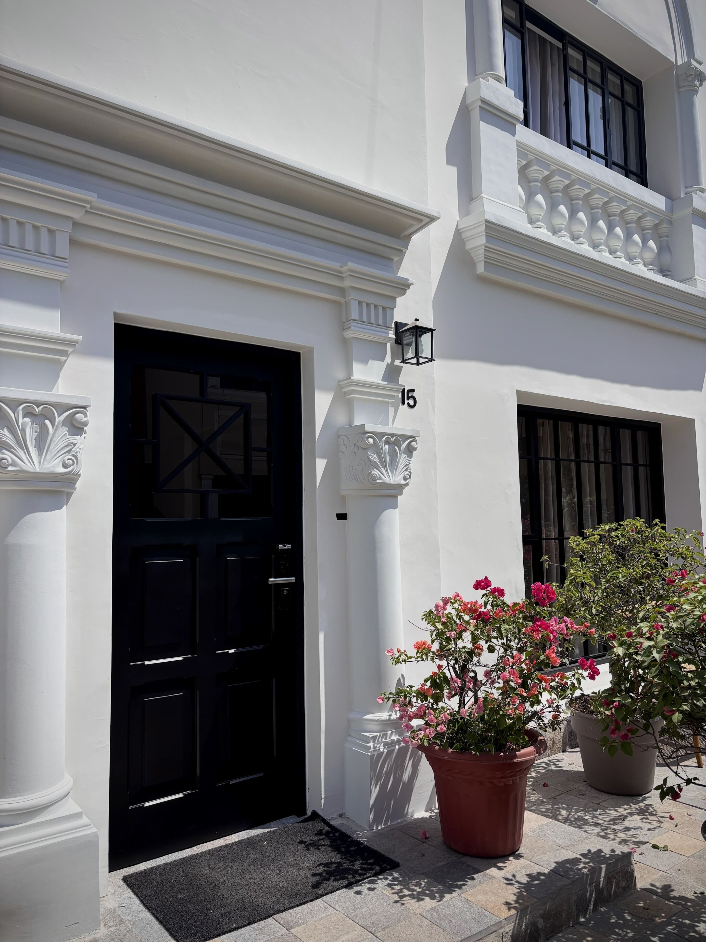 A white building with a black door, a number '15' on the wall, and potted plants with pink flowers.